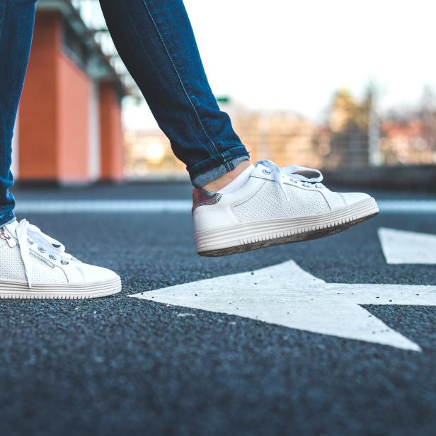 Eine Frau mit Jeans und weißen Turnschuhen geht auf einer Straße mit Richtungspfeil.