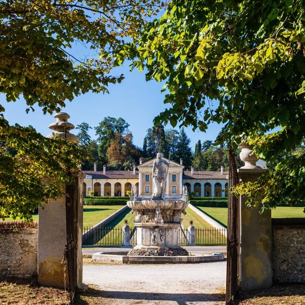 Blick durch ein offenes Tor auf eine Villa mit einem Brunnen im Garten.