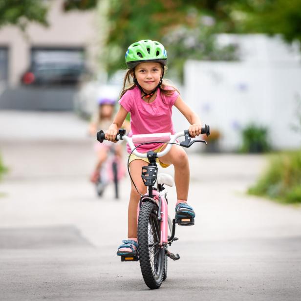 Ein Mädchen mit Helm fährt auf der Straße Fahrrad, ein weiteres Mädchen im Hintergrund.