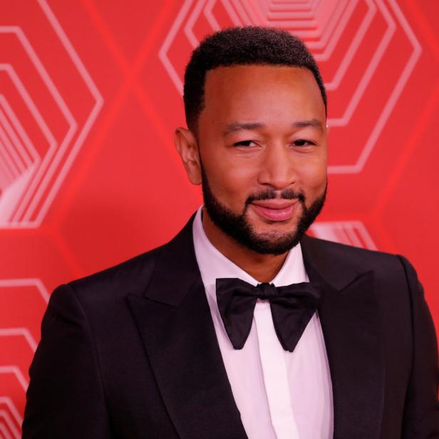 John Legend trägt einen Smoking mit Fliege vor einem roten Hintergrund.