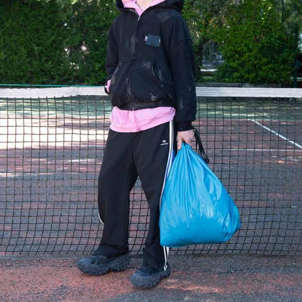Eine Person in Sportkleidung steht mit einer blauen Mülltüte auf einem Tennisplatz.