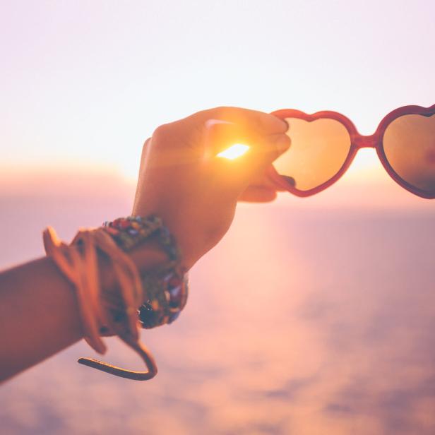Eine Hand hält eine herzförmige Sonnenbrille vor einem Sonnenuntergang.