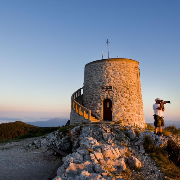 Ein Fotograf steht neben einem Steinturm auf einem Berggipfel und fotografiert die Landschaft.
