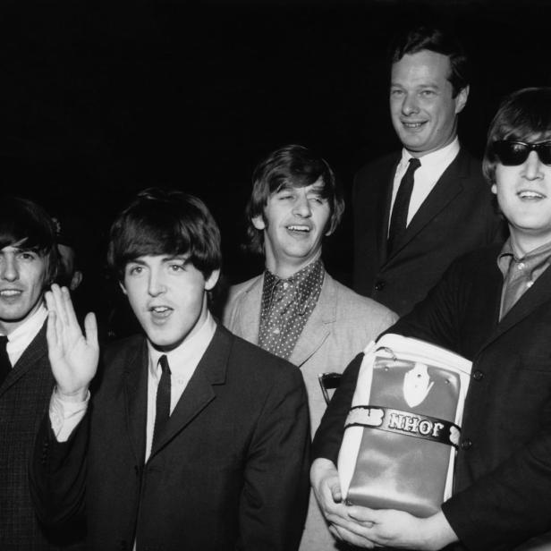 Die Beatles posieren mit einer Tasche mit der Aufschrift „John“ für ein Schwarzweißfoto.