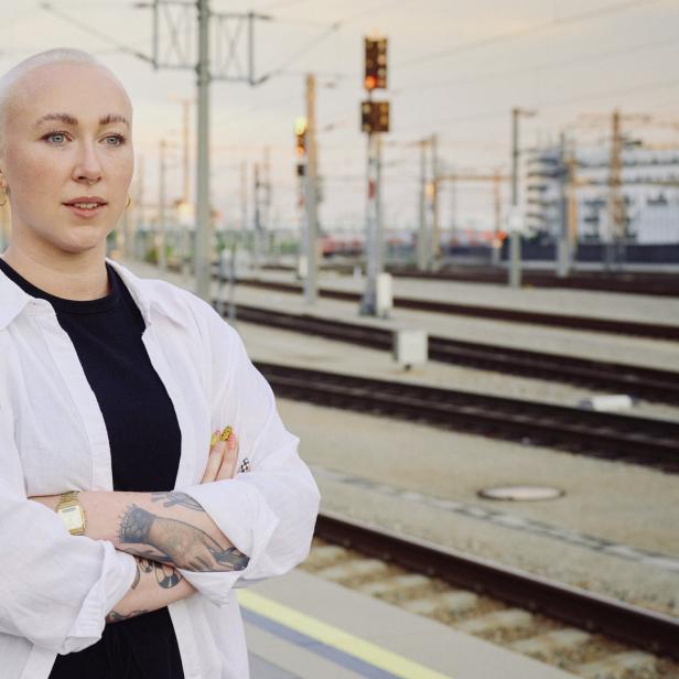 Eine Frau mit kurz geschorenen Haaren steht an einem Bahnhof.