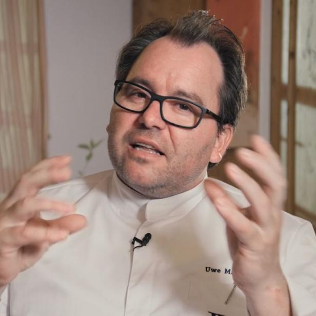 Uwe M., ein Mann mit Brille und Kochjacke, gestikuliert vor einem Fenster.