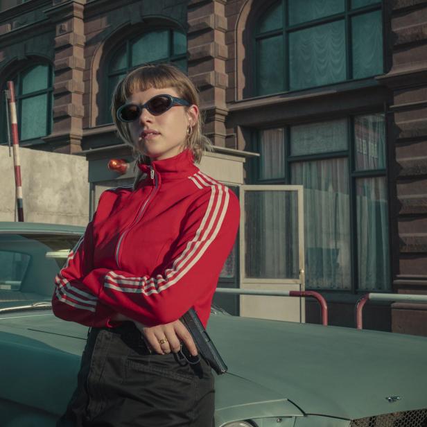 Eine Frau mit Sonnenbrille und roter Jacke lehnt an einem Oldtimer und hält eine Pistole.