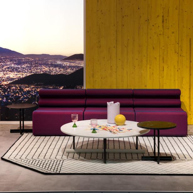 Ein lila Sofa, ein runder Tisch mit Gläsern und Mikado vor einer gelben Wand und einem Stadtbild bei Nacht.