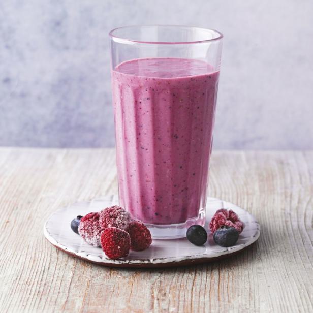 Ein Glas mit einem rosa Smoothie steht auf einem Teller mit gefrorenen Beeren.