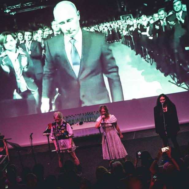 Eine Band tritt vor einer Leinwand mit einem Bild von Wladimir Putin auf.