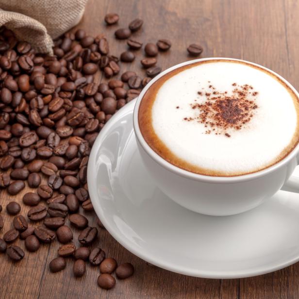 Eine Tasse Cappuccino steht auf einer Untertasse neben einem Sack Kaffeebohnen.