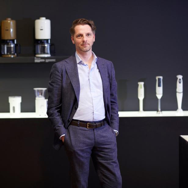 Ein Mann in einem Anzug steht vor einer Wand mit Kaffeemaschinen und Mixern.