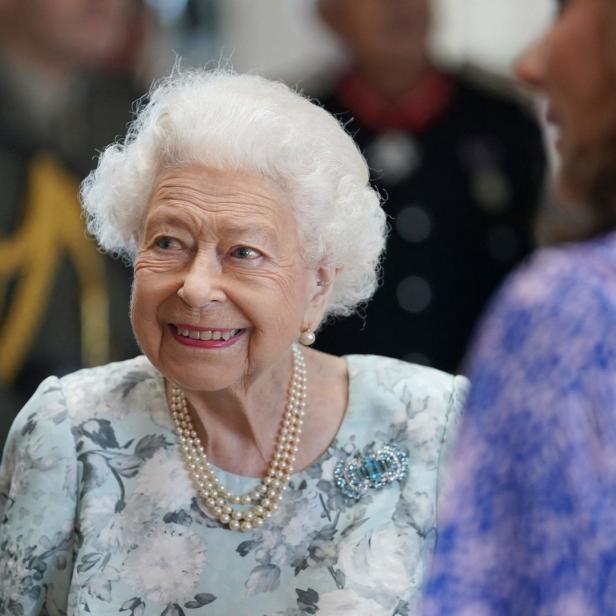 Königin Elizabeth II. lächelt in einem hellblauen Kleid mit Blumenmuster und einer Perlenkette.