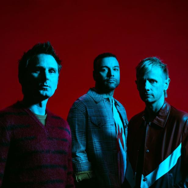Die Band Muse posiert vor einem roten Hintergrund.