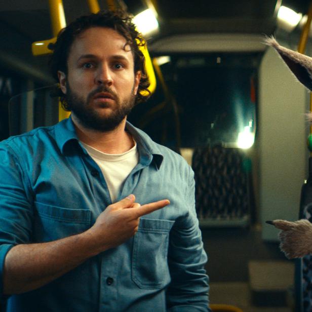Ein Mann und ein Känguru sitzen in einem Bus und zeigen mit dem Finger in die gleiche Richtung.