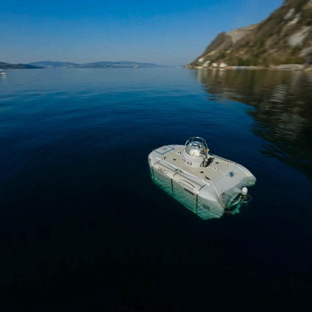 Ein kleines, weißes U-Boot fährt auf einem ruhigen See entlang einer bewaldeten Küste.