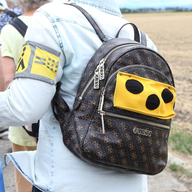 Person mit Rucksack und gelbem Armband mit Symbolen, im Freien auf einem Feldweg unterwegs.