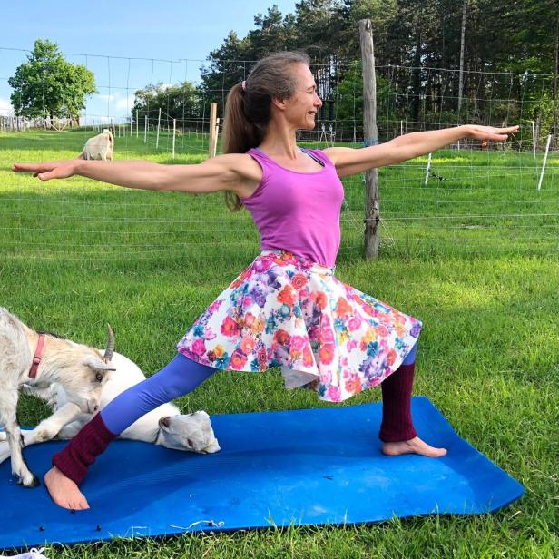 Eine Frau macht Yoga auf einer Matte, umgeben von Ziegen auf einer grünen Wiese.