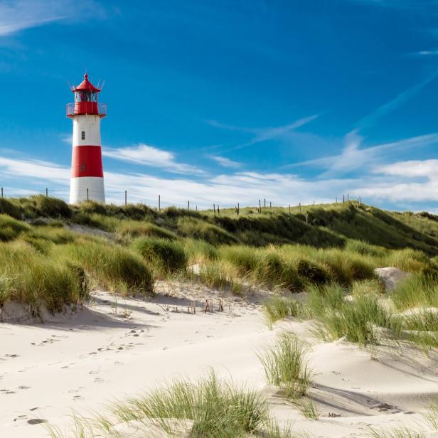 Ein rot-weiß gestreifter Leuchtturm auf einer Düne mit Strandhafer unter blauem Himmel.
