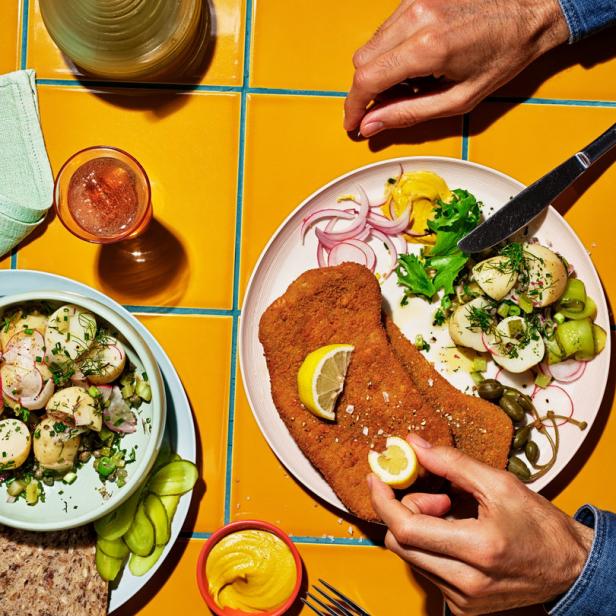 Ein Wiener Schnitzel wird mit Zitrone, Kartoffelsalat und Senf auf einem gelben Tisch serviert.