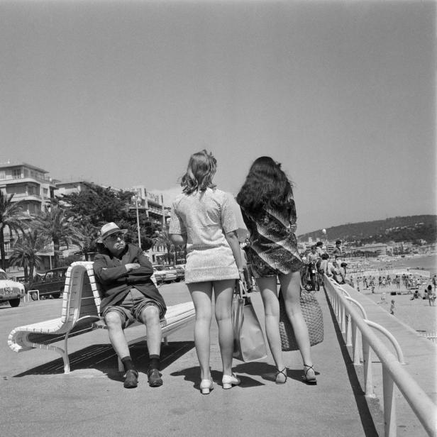 Ein Mann sitzt auf einer Bank, während zwei junge Frauen an der Promenade entlanggehen.