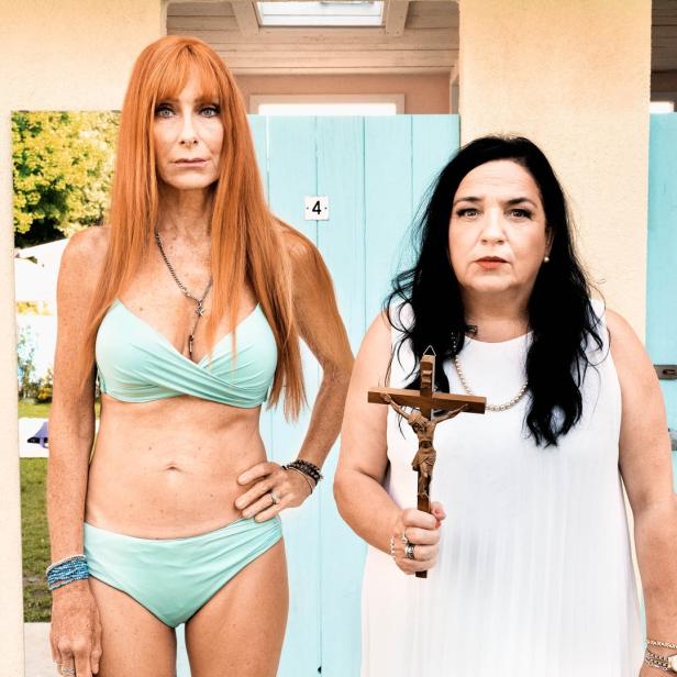 Zwei Frauen posieren vor hellblauen Strandkabinen, eine im Bikini, die andere mit einem Kruzifix.