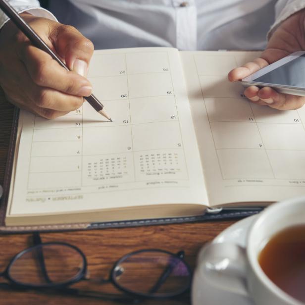 Eine Person schreibt mit einem Stift in einen Kalender und hält dabei ein Smartphone, auf dem Tisch liegen eine Brille und eine Tasse Tee.