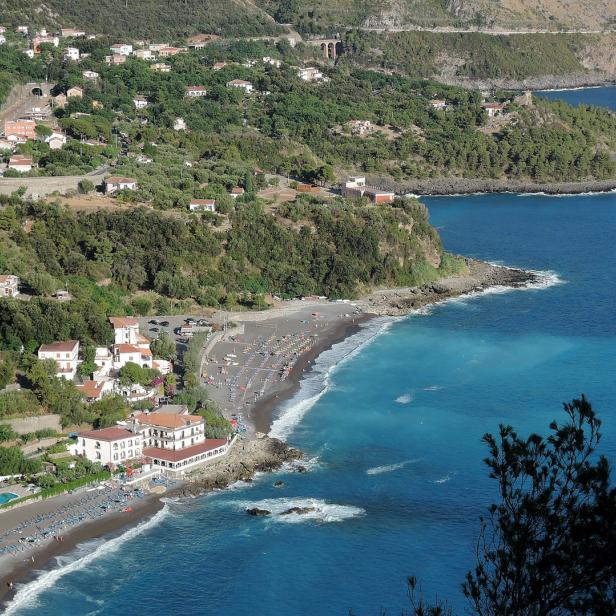 Blick auf einen Küstenort mit dunklem Sandstrand und türkisfarbenem Wasser.