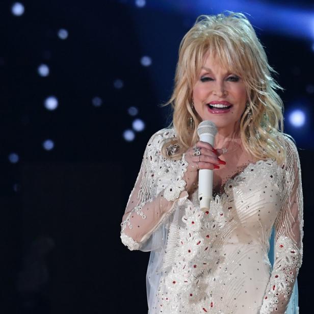 Dolly Parton singt in einem weißen, verzierten Kleid auf der Bühne.