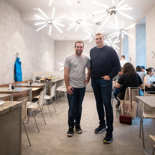 Zwei Männer stehen in einem hellen Restaurant mit Gästen an den Tischen.