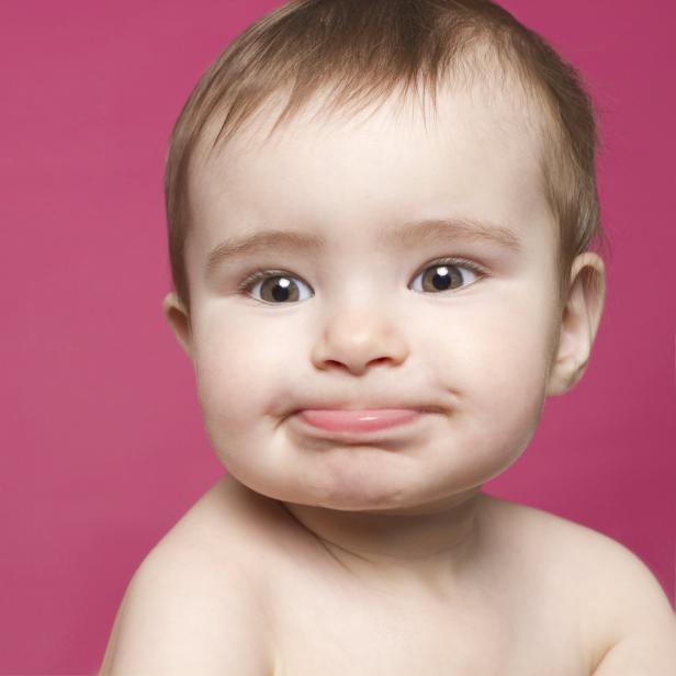 Ein Baby mit hellbraunen Haaren und großen Augen zieht eine Grimasse vor einem pinkfarbenen Hintergrund.