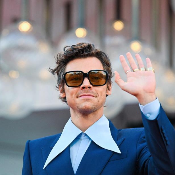 Harry Styles mit Sonnenbrille und blauem Anzug winkt in die Kamera.