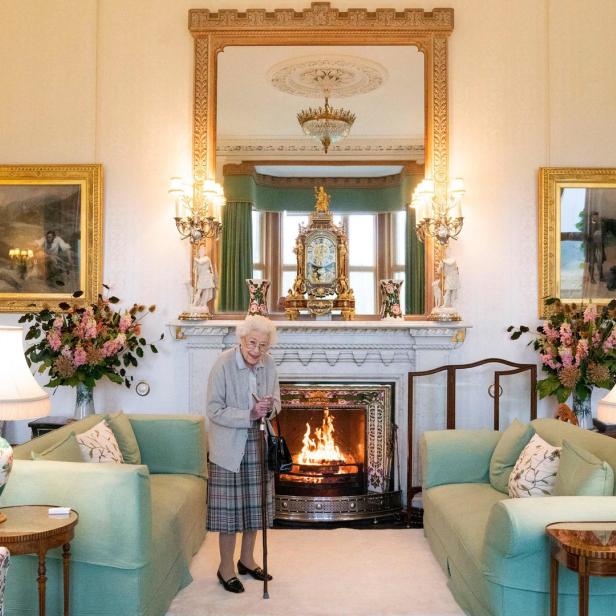 Queen Elizabeth II. steht mit einem Gehstock in einem eleganten Wohnzimmer vor einem Kamin.