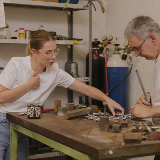 Eine junge Frau und ein Mann arbeiten an einem Tisch mit Metallteilen in einer Werkstatt.