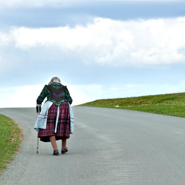 Eine ältere Frau mit Stock und traditioneller Kleidung geht allein eine Landstraße entlang.