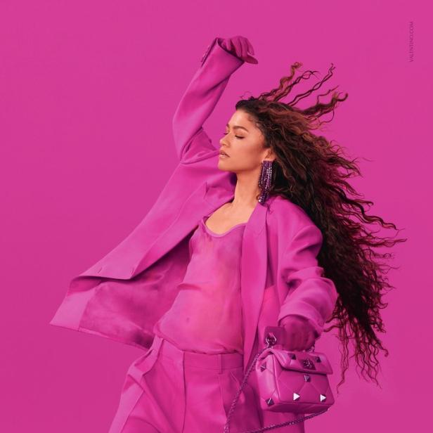 Zendaya trägt einen pinkfarbenen Hosenanzug und eine passende Handtasche vor einem pinkfarbenen Hintergrund.