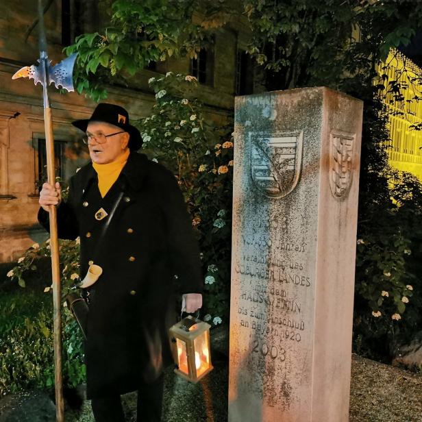 Ein Mann in historischer Kleidung steht mit einer Hellebarde und einer Laterne vor einem Denkmal.