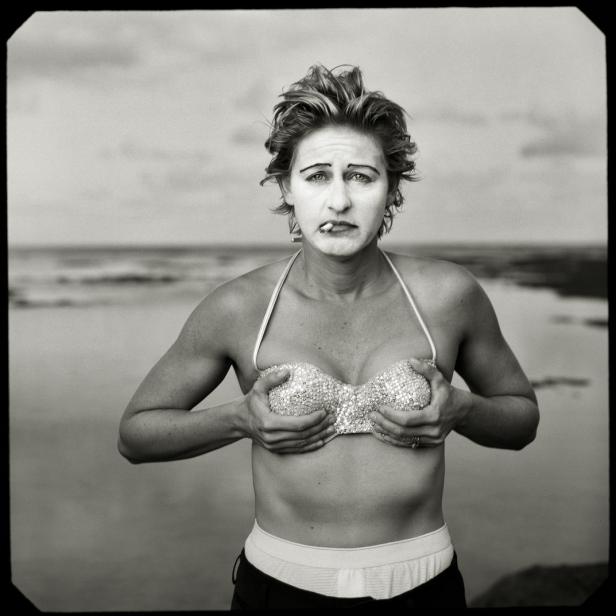 Eine Frau mit Clownsgesicht und glitzerndem Bikini-Oberteil am Strand raucht eine Zigarette.