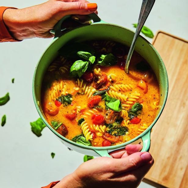 Hände halten einen Topf mit Tomatensuppe, Nudeln und Basilikum.