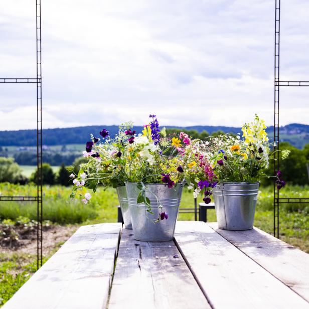 Drei Zinkeimer mit bunten Blumen stehen auf einem Holztisch im Freien.
