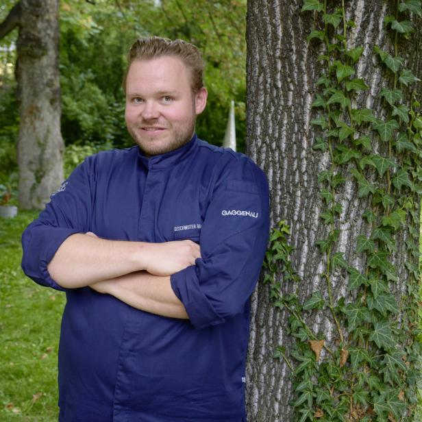 Ein Mann in Kochkleidung lehnt an einem Baum mit Efeu im Grünen.