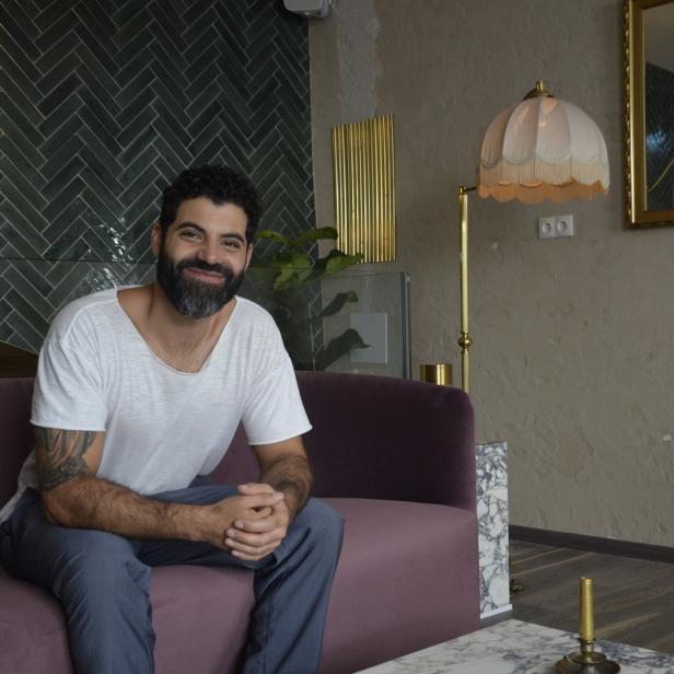 Ein lächelnder Mann mit Bart sitzt auf einem lila Sofa in einem stilvollen Raum.