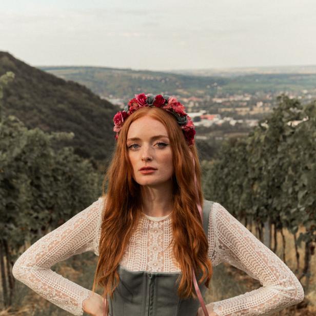 Eine Frau mit roten Haaren und Blumenkranz steht in einem Dirndl in den Weinbergen.