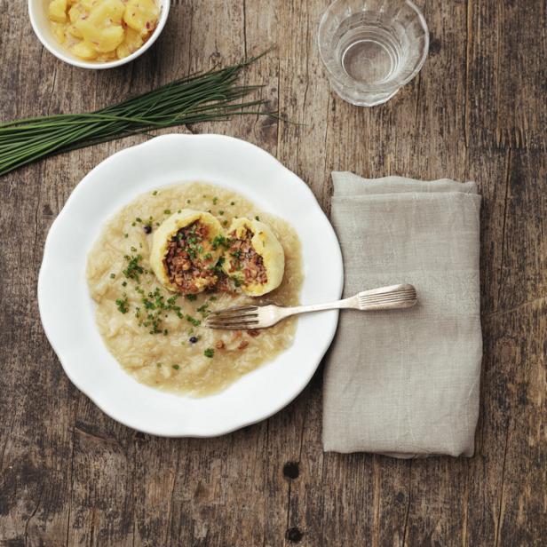 Ein Teller mit gefüllten Knödeln auf Sauerkraut, dazu Kartoffelsalat und Schnittlauch auf einem Holztisch.