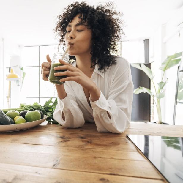 Eine Frau mit lockigem Haar trinkt einen grünen Smoothie in einer hellen Küche.