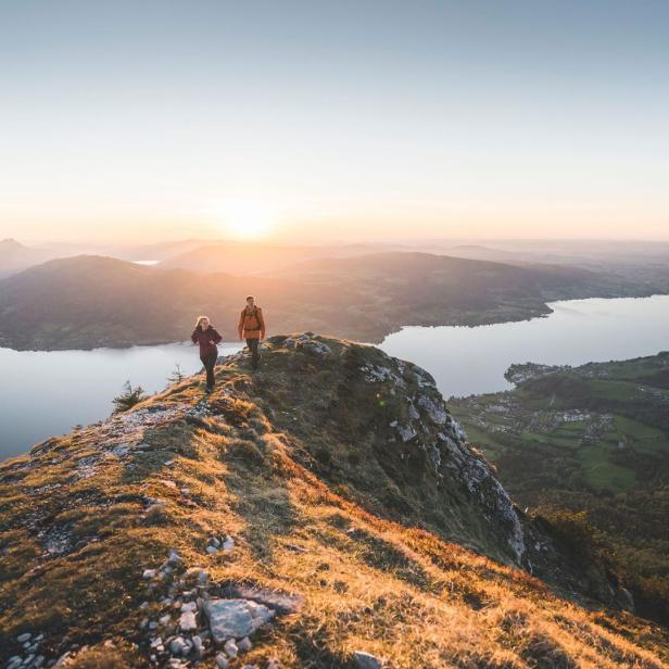 Zwei Wanderer auf einem Bergrücken mit Blick auf einen See bei Sonnenaufgang.