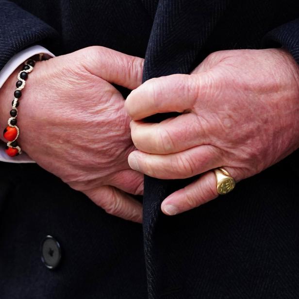 Die Hände einer Person mit einem Armband und einem goldenen Ring halten einen dunklen Mantel.