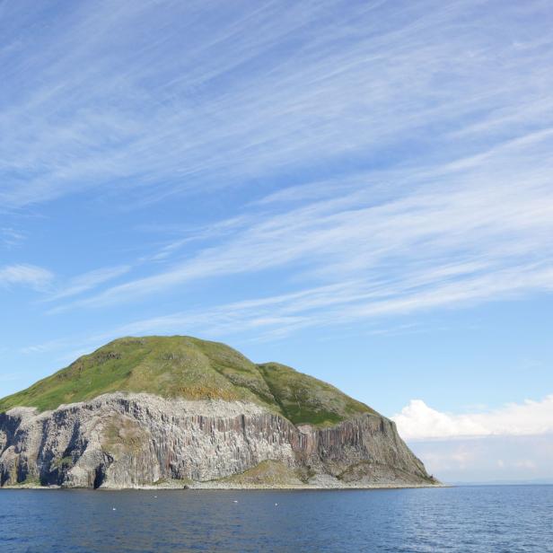 Ein kleiner, grüner Inselberg ragt aus dem blauen Meer unter einem hellen Himmel.