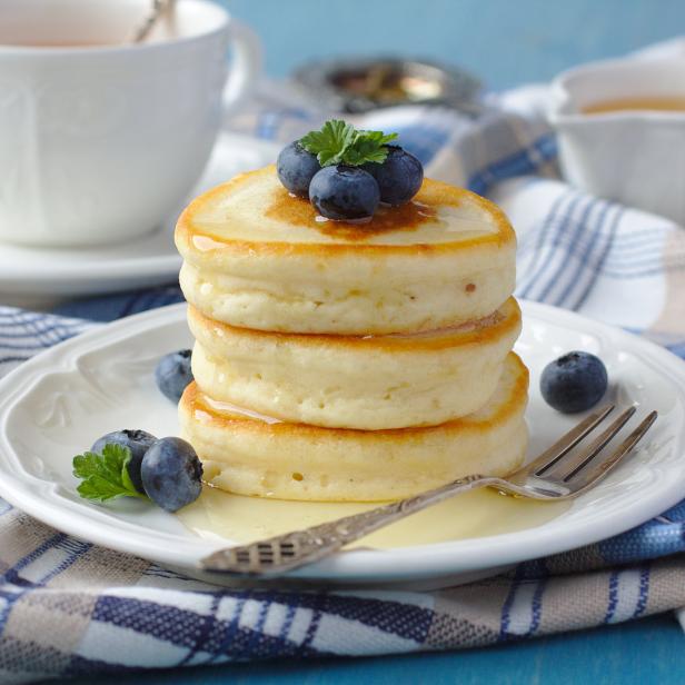 Ein Stapel Pfannkuchen mit Blaubeeren und Sirup auf einem Teller neben einer Tasse Tee.