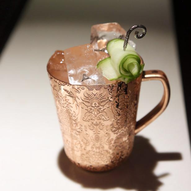 Ein Cocktail im Kupferbecher mit Eis und Gurkengarnitur steht auf einem Tisch.